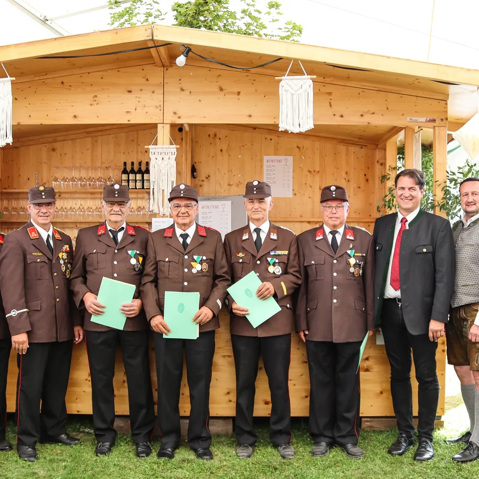 Sechs Männer in braunen Uniformen mit Medaillen stehen vor einem hölzernen Stand und halten grüne Papiere. Sie werden von einem lächelnden Mann in Anzug und zwei anderen in Freizeitkleidung umgeben.
