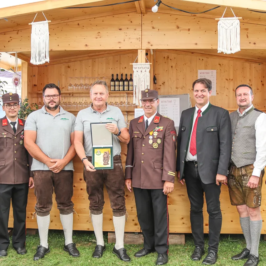 Eine Gruppe von Männern in Uniformen und traditioneller Kleidung steht vor einer Holzbar, einer Mann hält eine Auszeichnung.