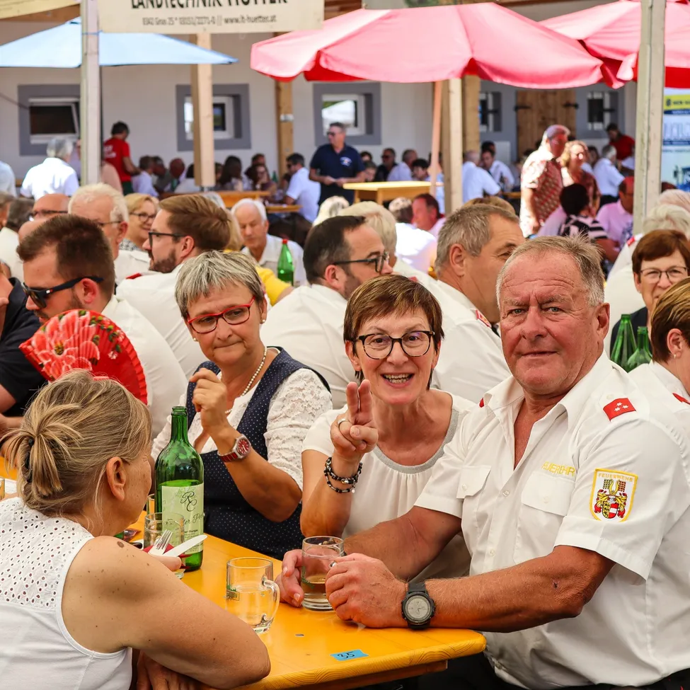 Eine Gruppe von Menschen sitzt an einem Tisch in einem Restaurant im Freien, einige tragen Brillen und Uniformen. Der Mann in der Mitte macht ein Friedenszeichen.