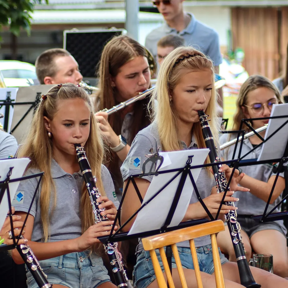 Eine Gruppe junger Musiker, darunter Mädchen, die Klarinetten und Flöten spielen, tritt im Freien auf. Sie sind lässig gekleidet und scheinen sich auf ihre Musik zu konzentrieren.