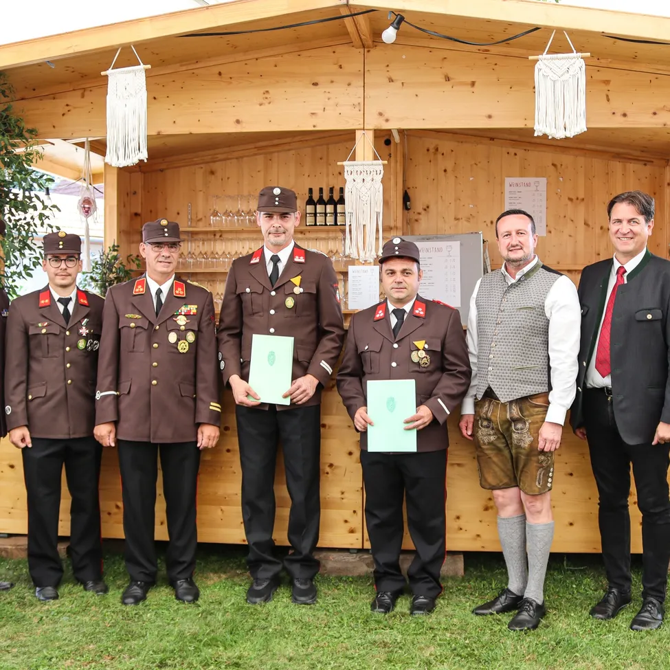 Eine Gruppe von Männern in Uniform, die Urkunden halten, steht vor einer Holzhütte mit Flaschen und Dekorationen.
