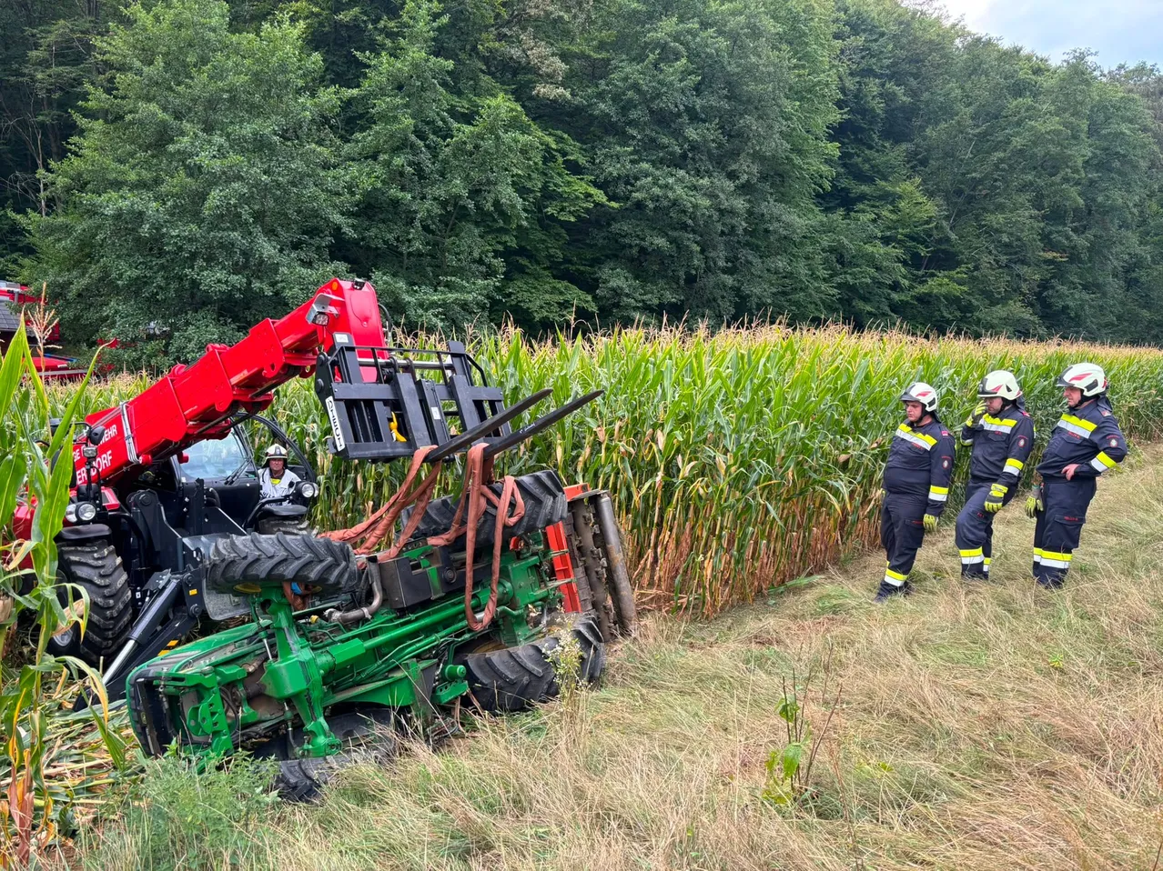 Ein grüner Traktor liegt umgedreht in einem Maisfeld, Feuerwehrleute in Uniform sind in der Nähe.