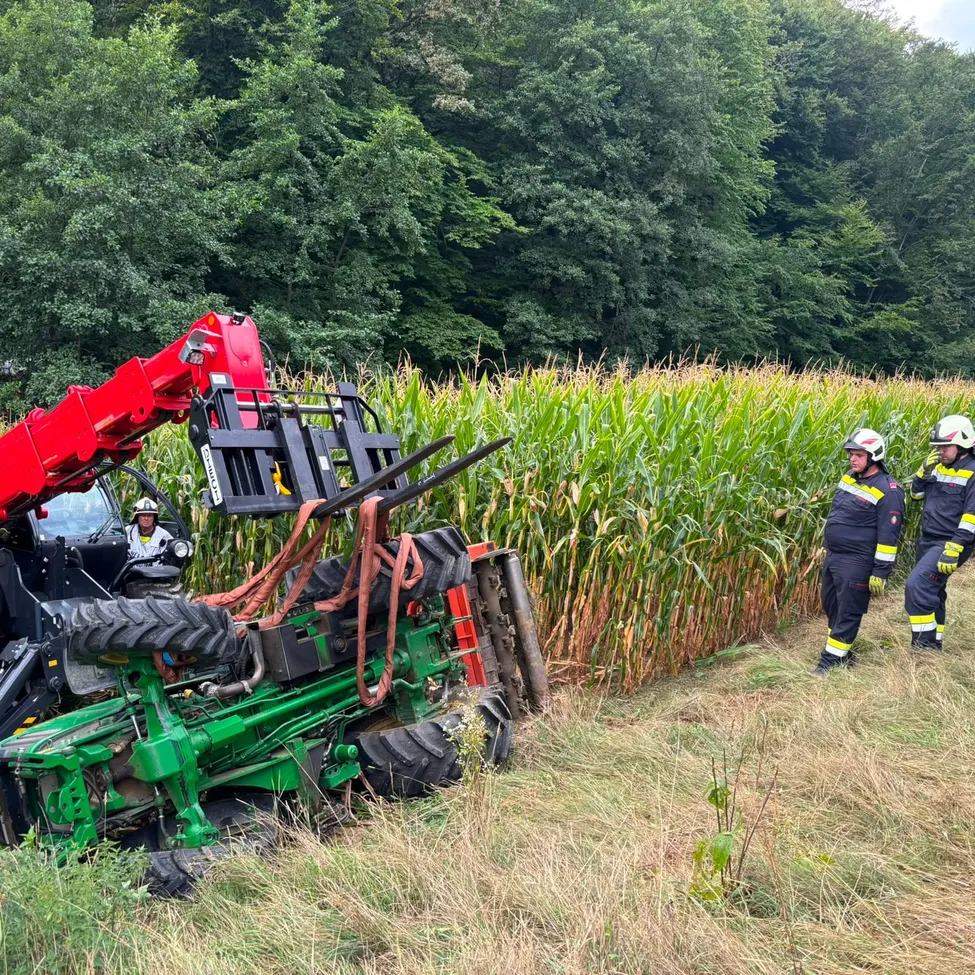 Ein grüner Traktor liegt umgedreht in einem Maisfeld, Feuerwehrleute in Uniform sind in der Nähe.