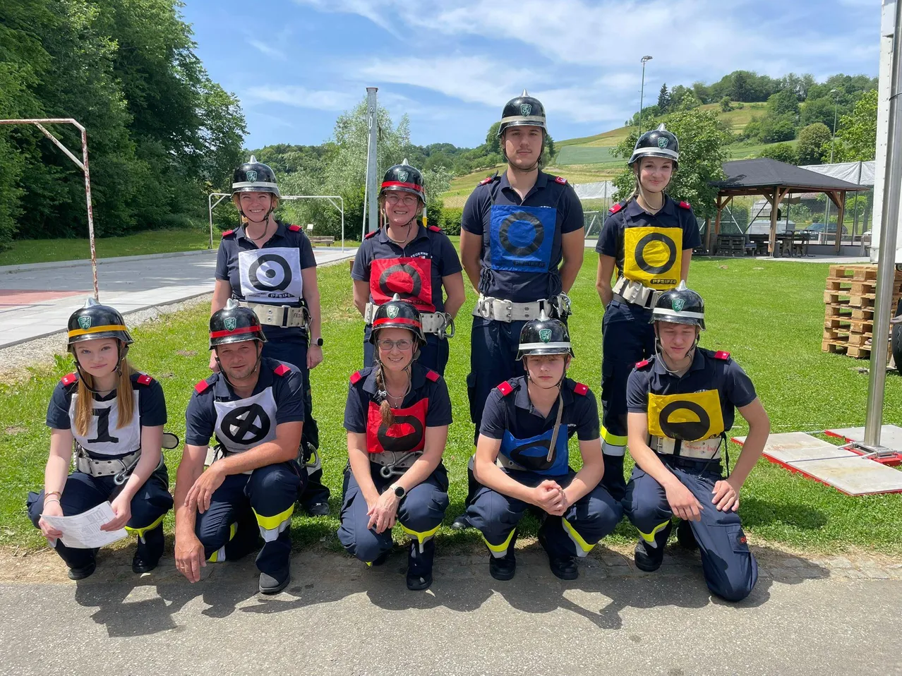 Eine Gruppe von Menschen in Uniformen und Helmen posiert für ein Foto auf einem Rasenfeld mit Bäumen und einem Pavillon im Hintergrund.