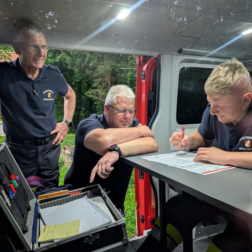 Three men in uniforms, possibly firefighters, are inside a van. One is writing on a paper while the other two are looking at him.