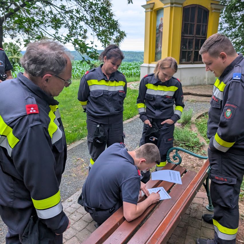 Five firefighters are gathered outdoors, three standing and two seated. One seated firefighter is writing on a piece of paper. They are in front of a yellow building with a statue and surrounded by greenery.