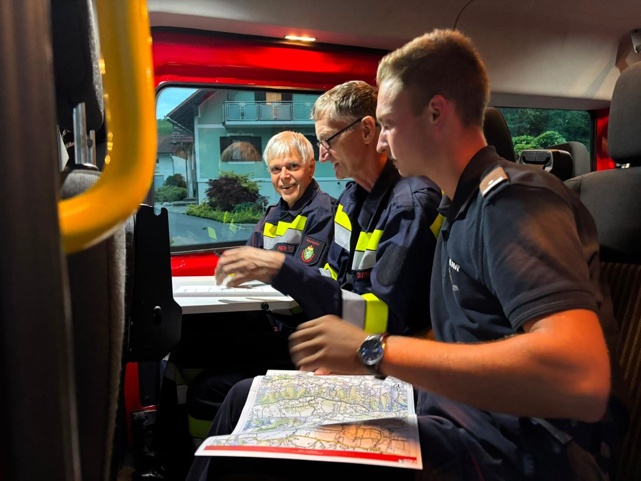 Three firefighters sit inside a vehicle, two are smiling, and one is holding a map. A house is visible through the window.