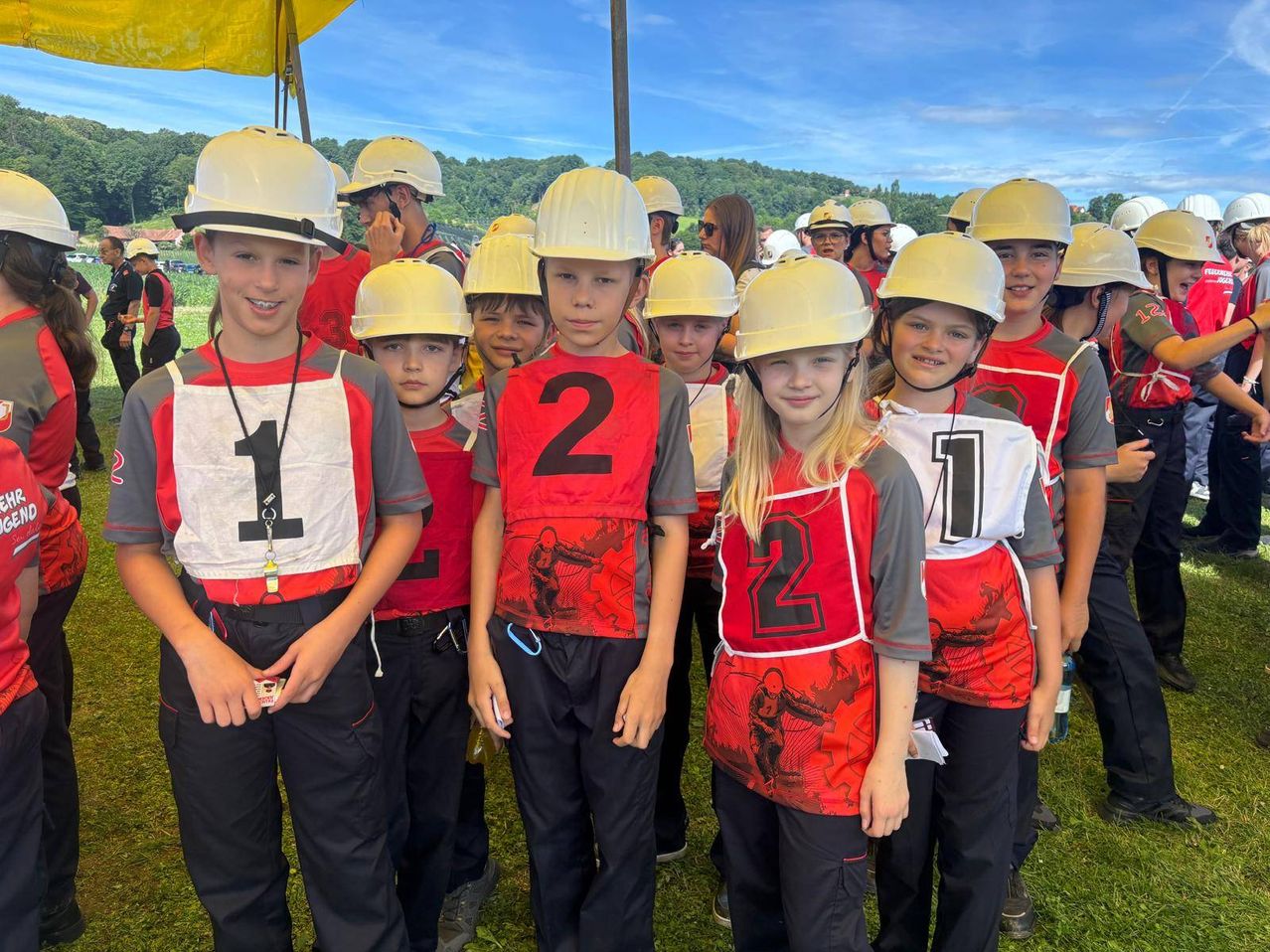 A group of young athletes in red and gray uniforms, with numbers on their vests, stand in a field under a clear blue sky.