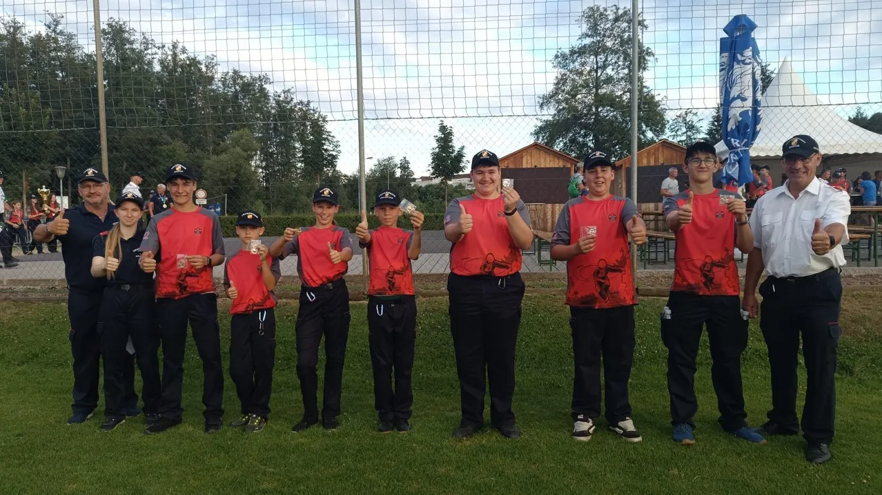 Sechs junge Athleten in roten Trikots posieren für ein Foto auf einem Feld und halten kleine Karten in der Hand. Dahinter ist ein Netz und Bäume zu sehen.