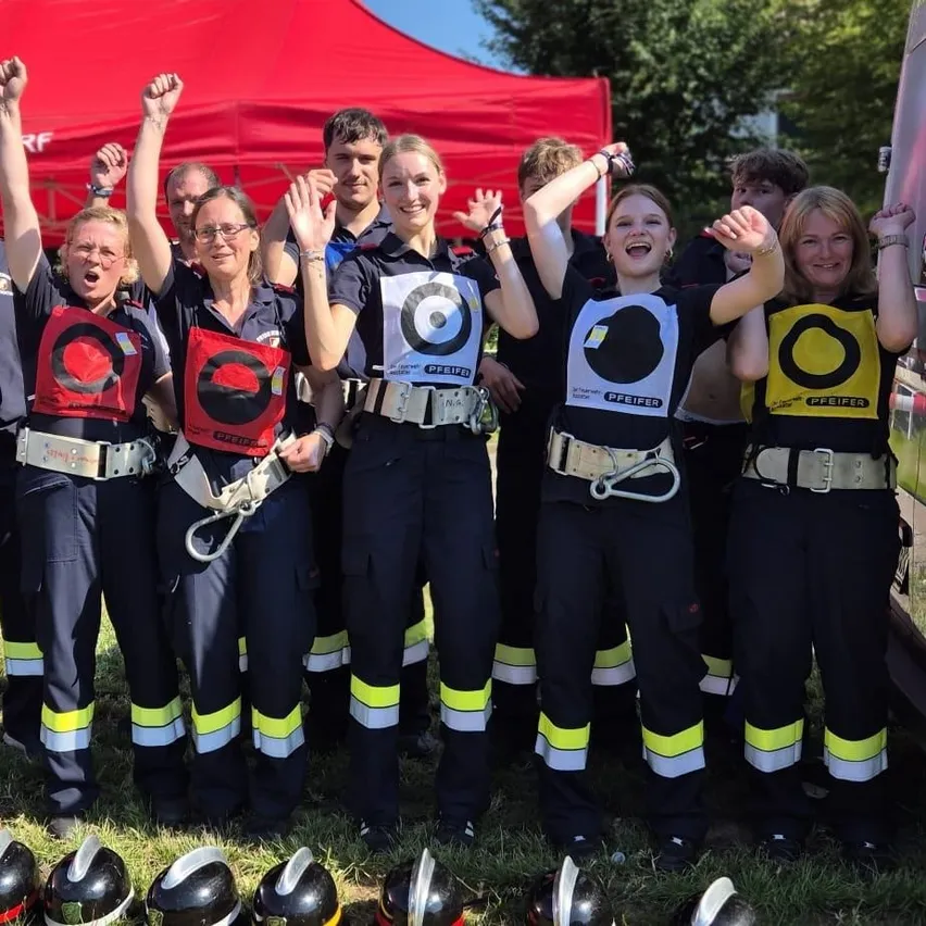 Eine Gruppe von Menschen in Sicherheitsuniformen steht unter einem roten Zelt. Sie posieren für ein Foto mit erhobenen Armen. Sie stehen vor einem Feuerwehrwagen.