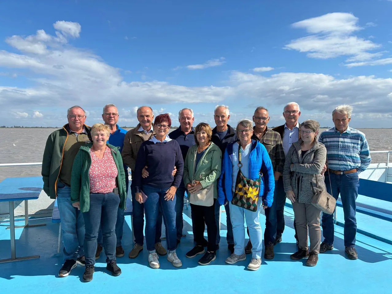 Eine Gruppe von Menschen, darunter Männer und Frauen, steht zusammen auf einer blauen Plattform, möglicherweise auf einem Boot, unter einem blauen Himmel mit Wolken.