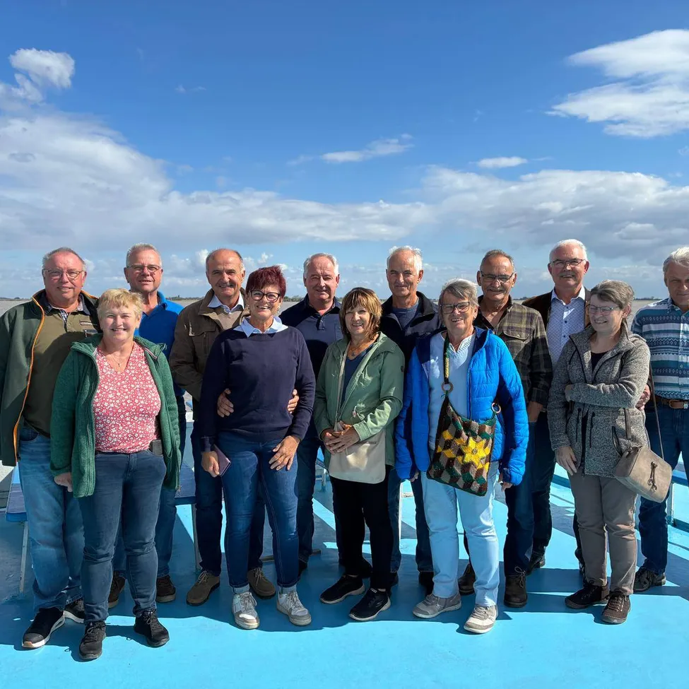 Eine Gruppe von Menschen, darunter Männer und Frauen, steht zusammen auf einer blauen Plattform, möglicherweise auf einem Boot, unter einem blauen Himmel mit Wolken.