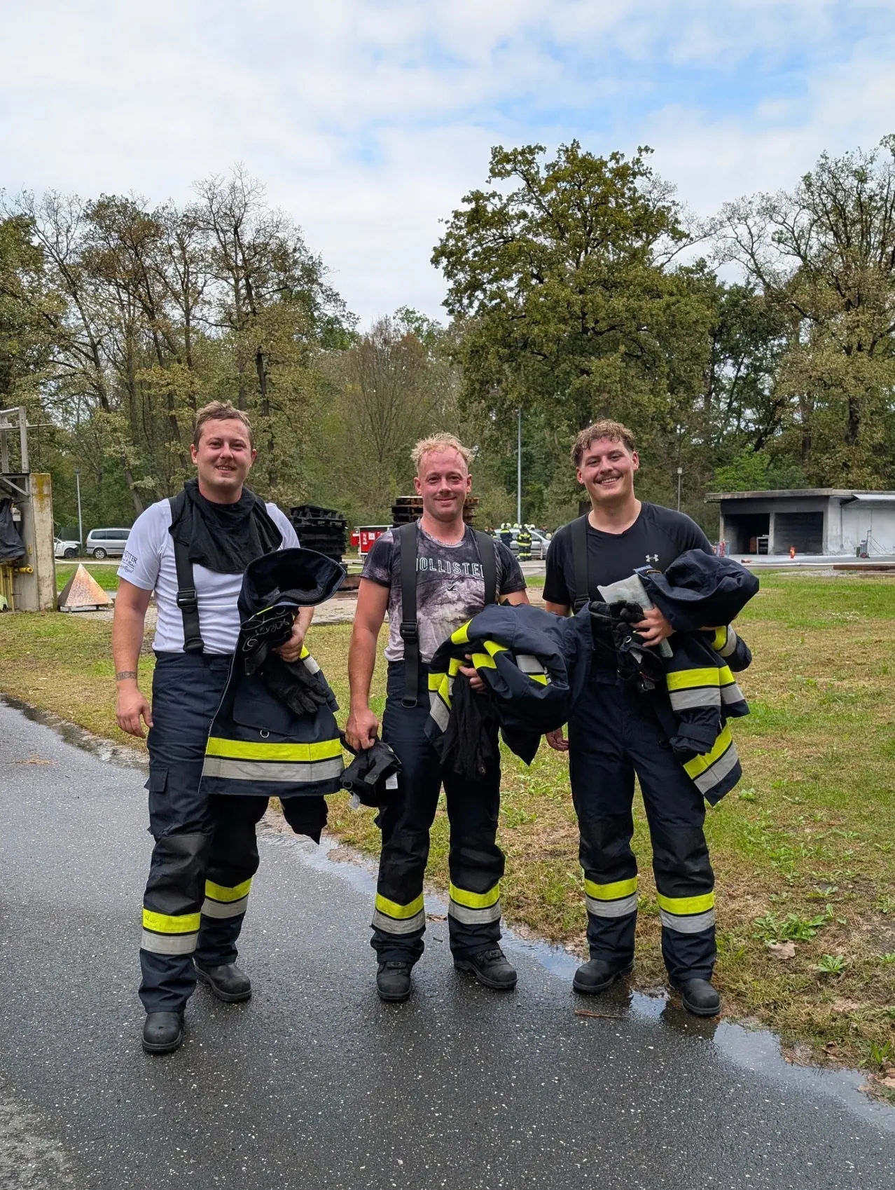 Drei Feuerwehrleute stehen auf einer nassen Straße, lächeln und halten ihre Uniformen. Dahinter ein Rasenbereich mit Bäumen und einem Gebäude.