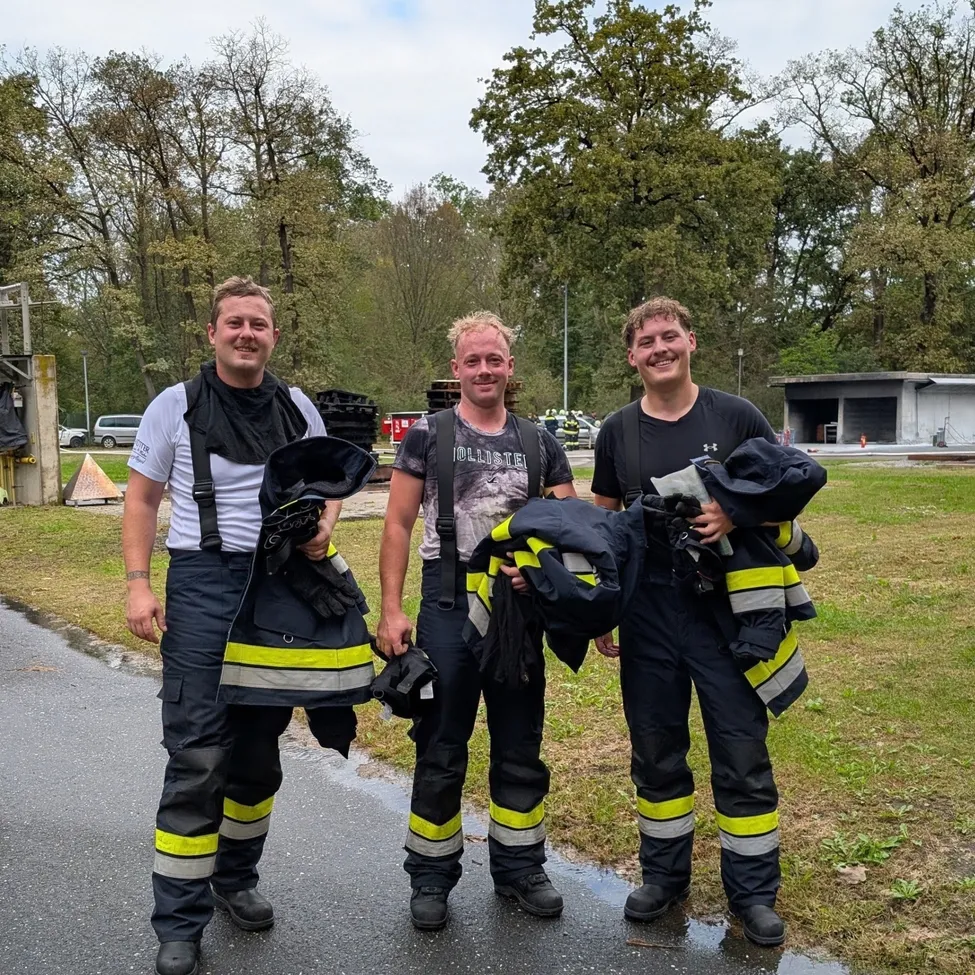Drei Feuerwehrleute stehen auf einer nassen Straße, lächeln und halten ihre Uniformen. Dahinter ein Rasenbereich mit Bäumen und einem Gebäude.