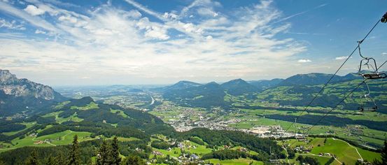 Luftaufnahme einer Bergregion mit üppigen grünen Tälern, einer kurvenreichen Straße und einem blauen Himmel, der von wolkenartigen Streifen durchzogen ist.