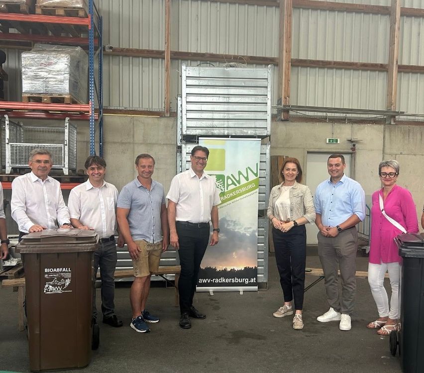 Eine Gruppe von Personen, die formell gekleidet sind, steht gemeinsam vor einem Banner. Der Hintergrund scheint ein Lager zu sein, mit Lagerboxen und einer Metallstruktur.