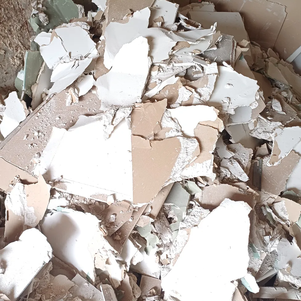 Ein Haufen zerbrochener Stücke von Gipskarton und Pappe liegt auf dem Boden verstreut, mit etwas Staub und Schmutz.