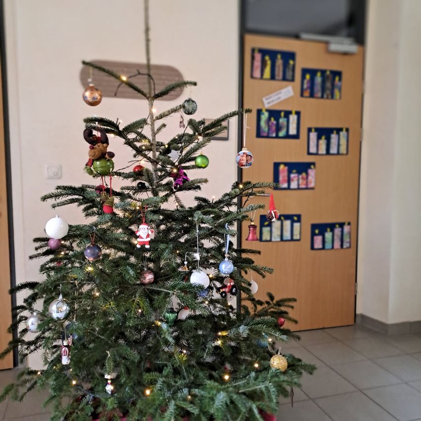 Ein Weihnachtsbaum mit bunten Dekorationen steht in einem Raum mit einer Holztür. Der Baum ist mit Lichtern und verschiedenen Ornamenten verziert. Die Tür ist braun und hat Bilder, die daran befestigt sind.
