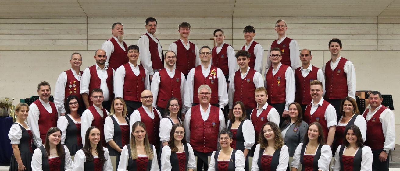 Ein Chor, gekleidet in passende rote und weiße Westen mit Medaillen um den Hals, posiert für ein Foto mit weißem Hintergrund.