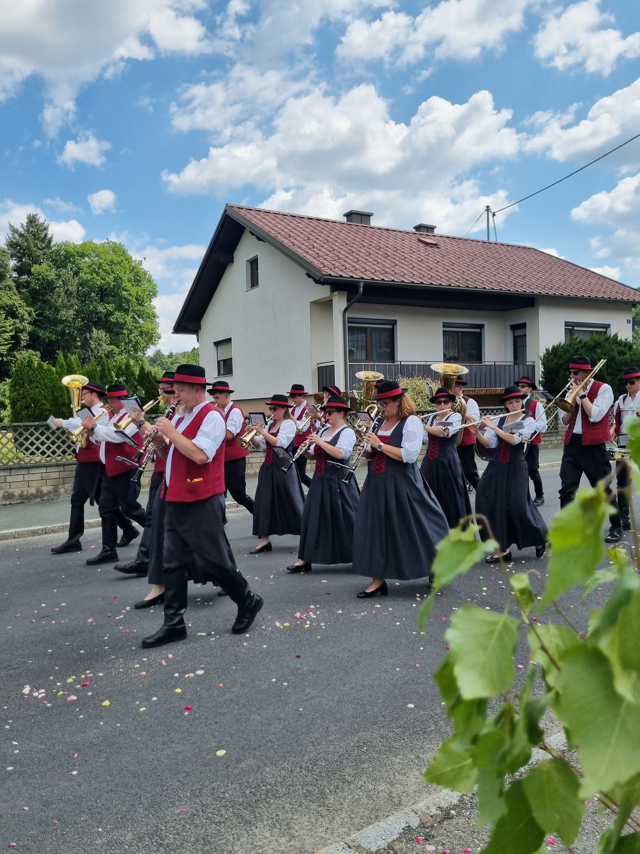 Eine Blaskapelle spielt auf der Straße, wobei die Musiker verschiedene Instrumente spielen. Sie sind in traditioneller Kleidung gekleidet und marschieren einreihig. Hinter ihnen steht ein Haus mit einem braunen Dach.
