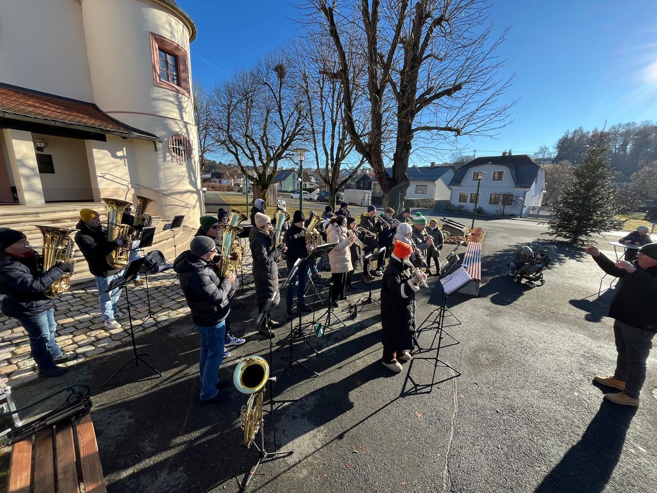 Eine Gruppe von Musikern in Winterkleidung spielt Instrumente im Freien. Im Hintergrund sind Bäume und Gebäude zu sehen.