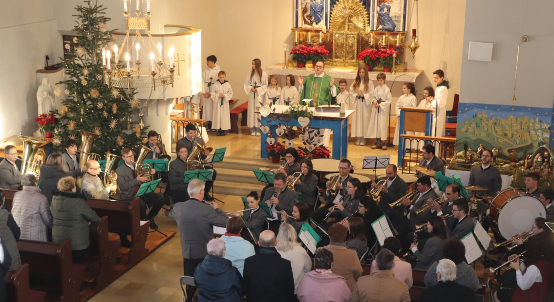 Ein Weihnachtschor und ein Blechbläserensemble spielen in einer Kirche. Der Chor steht hinter einem Priester am Altar, während das Blechbläserensemble auf dem Kirchenboden spielt. Das Publikum sitzt davor.