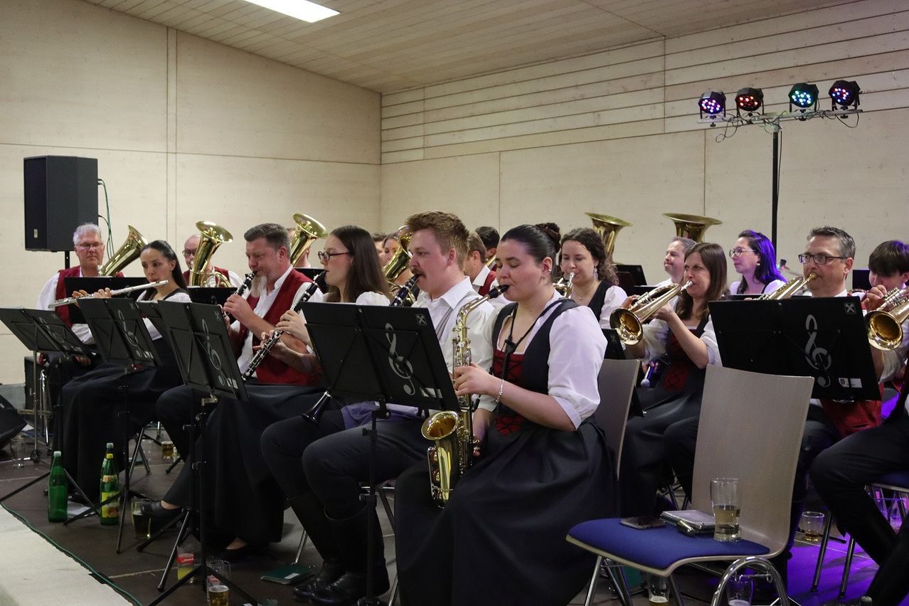 A band is seated in a room, performing on various brass and woodwind instruments. Some members are playing trumpets, others saxophones and clarinets. Glasses, drinks, and music stands are scattered around.