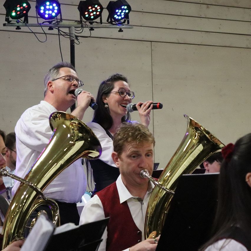 A band with two singers, a man and a woman, performs on stage with a tuba and microphones. The man wears a vest, and the woman wears glasses. They are illuminated by stage lights.