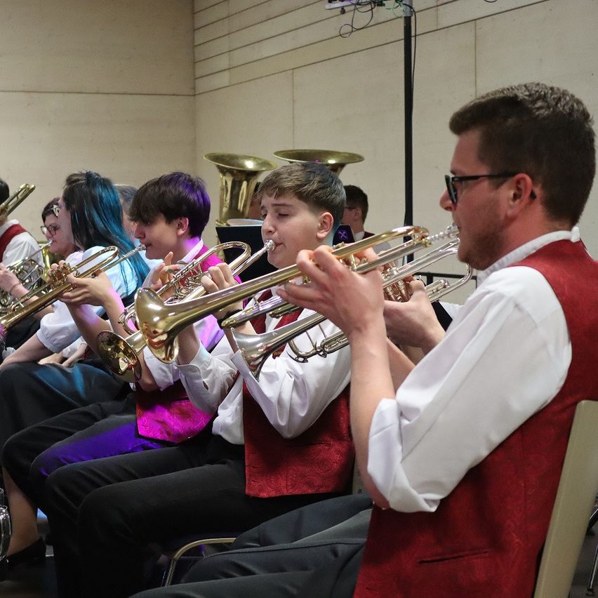 A group of young musicians wearing red vests are playing trumpets. They are seated in a row, with one player at the front. Behind them, other musicians play various instruments.