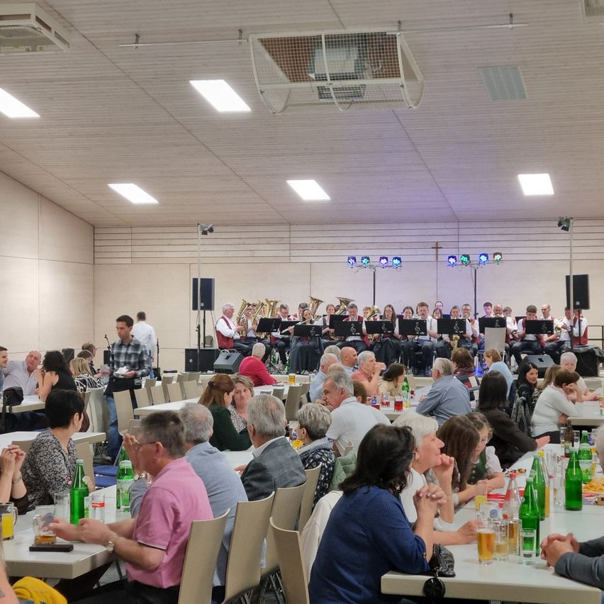 A large gathering of people, seated and standing, in a room. A band plays on stage in front of them. The room has white walls, ceiling lights, and tables with drinks and food.