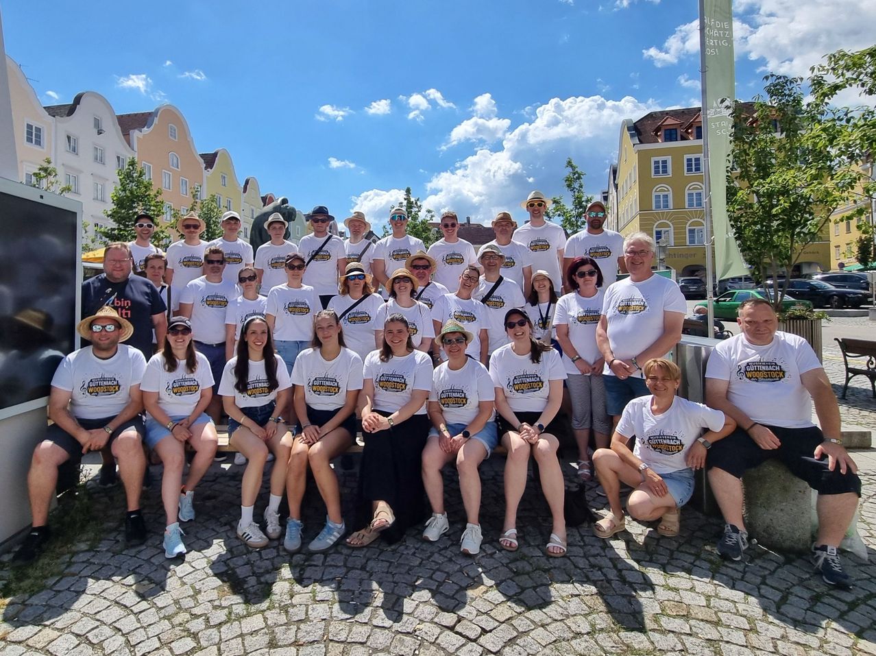 Eine Gruppe von Menschen in weißen T-Shirts mit aufgedruckten Designs und Hüten posiert für ein Foto im Freien. Einige sitzen auf einer Bank. Dahinter befinden sich Gebäude, Bäume und ein klarer blauer Himmel.