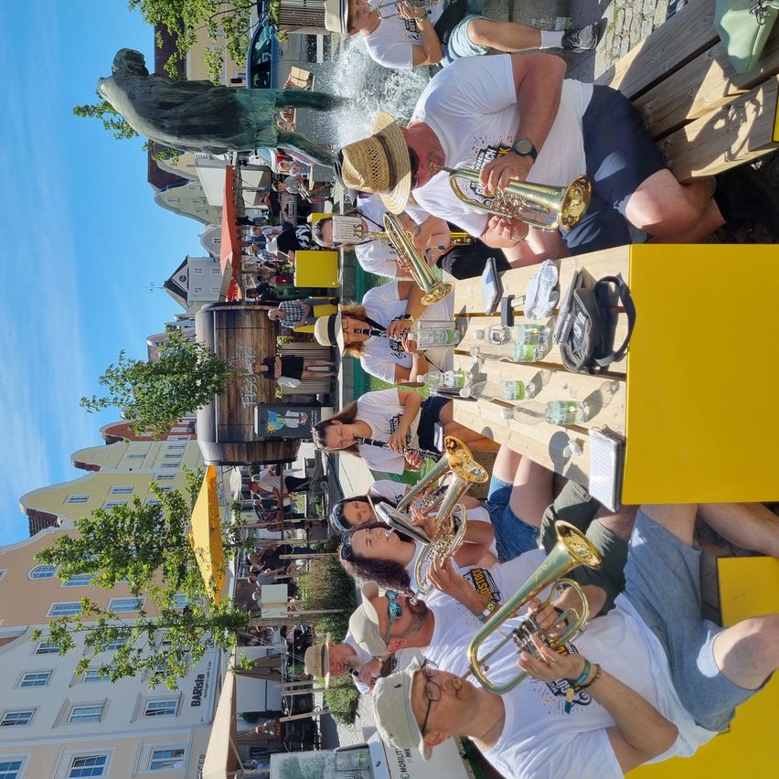 Eine Gruppe von Menschen sitzt auf einer Bank und spielt Trompeten. Einige tragen Hüte und Sonnenbrillen. Eine gelbe Box ist auf dem Tisch. Im Hintergrund befinden sich Gebäude, Bäume und Autos.
