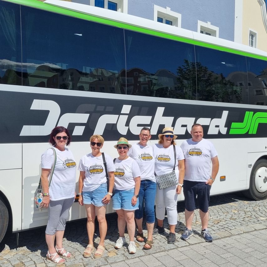 Eine Gruppe von sechs Personen, die weiße T-Shirts mit dem Wort Woodstock tragen, steht vor einem Bus. Sie alle lächeln und posieren für ein Foto.