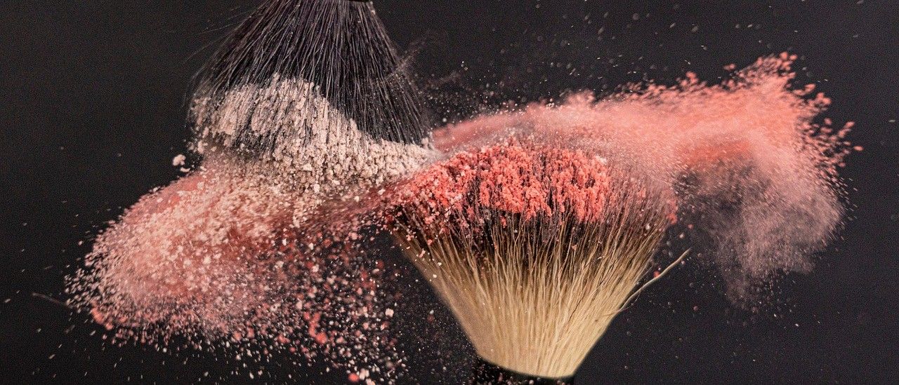 Ein Make-up-Pinsel mit rosa Puder darum herum, auf einem dunklen Hintergrund. Der Pinsel hat einen schwarzen Griff und eine weiße Borste. Das Puder ist rosa und scheint vom Pinsel zu fallen.
