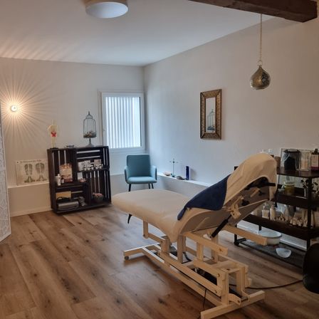 Ein gut beleuchteter Raum mit Holzboden, weißen Wänden und einem Massagetisch. Ein Regal, ein Stuhl und ein gerahmtes Bild sind an der Wand. Ein Schrank mit Gegenständen und einem Mannequin ist links aufgestellt.