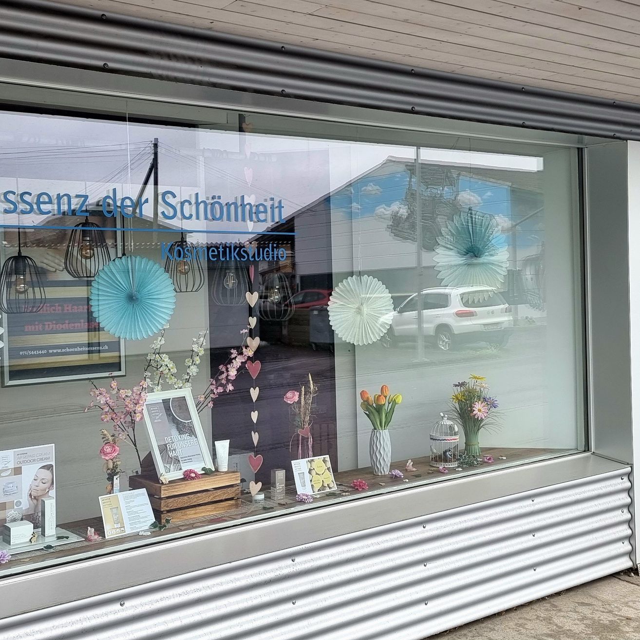 Ein Schaufenster zeigt verschiedene Dekorationsartikel wie Blumen, Bilderrahmen und dekorative Kugeln. Es gibt auch ein Schild mit der Aufschrift 'Ssenz der Schönheit Kosmetikstudio' und eine Reflexion eines Autos im Glas.
