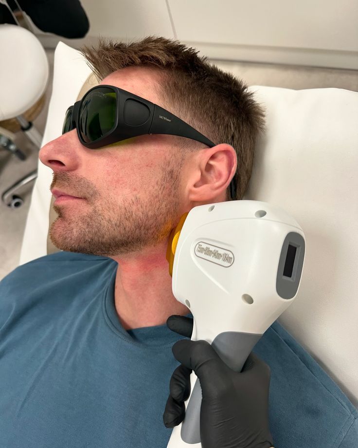 A man wearing protective glasses is lying on a pillow while undergoing a laser treatment on his neck.
