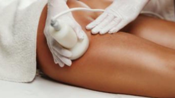 Eine Person in Handschuhen bekommt eine Beinmassage mit einem Gerät, während sie auf einem Bett liegt, teilweise von einem Handtuch bedeckt.