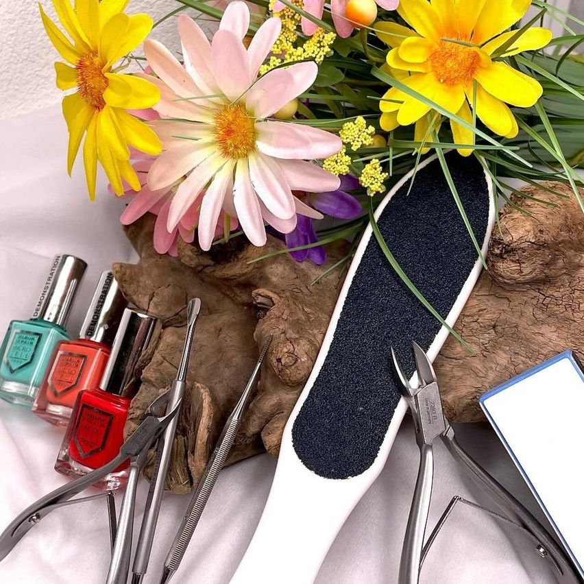 Ein Set von Nagelpflegewerkzeugen mit verschiedenen Instrumenten wie einer Fußfeile, Nagelknipsern und einem Nagelhautstäbchen. Die Werkzeuge liegen auf einer weißen Oberfläche, mit bunten Nagellackflaschen und Blumen im Hintergrund.