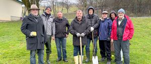 Eine Gruppe von Männern in Winterkleidung steht in einem Feld mit Schaufeln und bereitet wahrscheinlich den Boden für das Pflanzen vor. Bäume und ein Zaun sind im Hintergrund zu sehen.