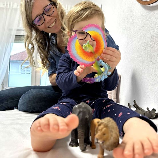 Eine Frau und ein Kind sitzen auf einem Bett mit Spielzeug um sie herum. Das Kind hält ein buntes Spielzeug, während die Frau lächelt und es ansieht.