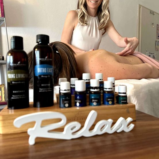 Eine Frau erhält eine Rückenmassage mit ätherischen Ölen. Ein Holzbrett mit dem Wort 'Relax' steht auf dem Tisch.
