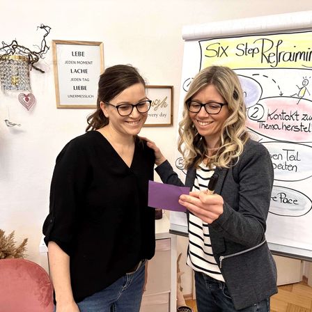 Zwei Frauen stehen neben einer Whiteboard mit motivierenden Texten und lächeln. Eine hält ein lila Papier und die andere schreibt darauf. Dahinter hat eine Wand einen gerahmten Zitat und dekorative Gegenstände.