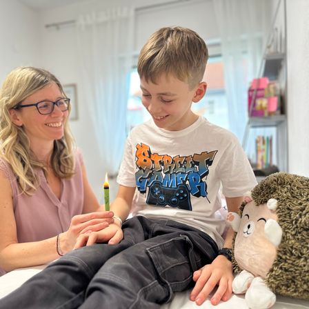 Ein lächelnder Junge mit einer brennenden Kerze sitzt auf einem Bett mit einer Frau, beide halten Händchen. Ein Igel-Plüschtier ist in der Nähe. Der Raum hat weiße Wände und Regale mit Büchern.