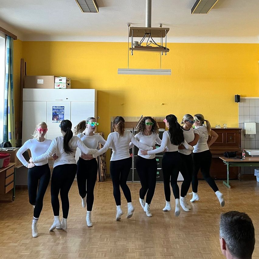 A group of women in matching outfits dance in a room with yellow walls and wooden floors, while a man watches from the side.