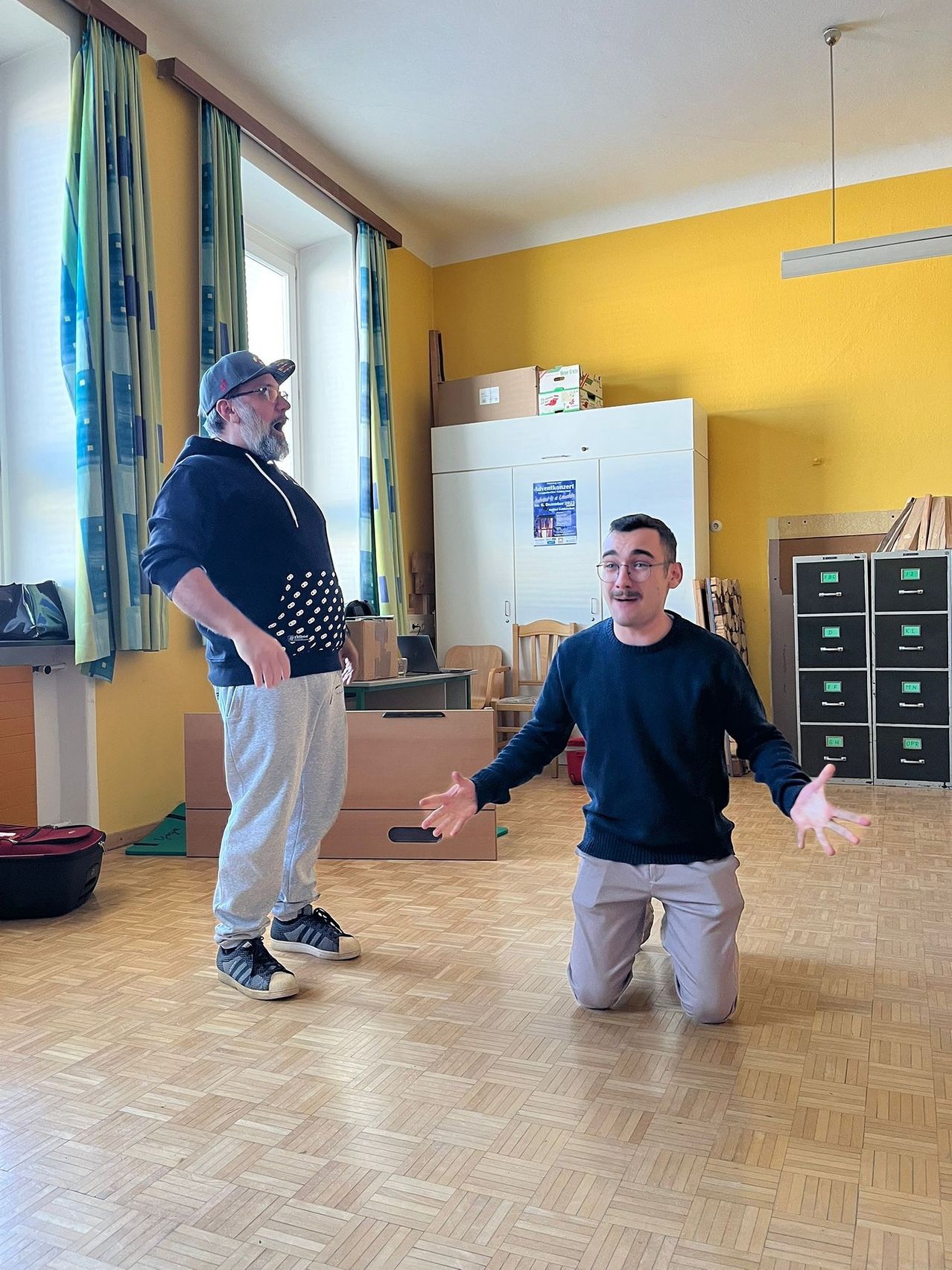 Two men in a room. One is kneeling and the other is standing. They are wearing casual clothes. There are cabinets, a desk, and a window behind them.
