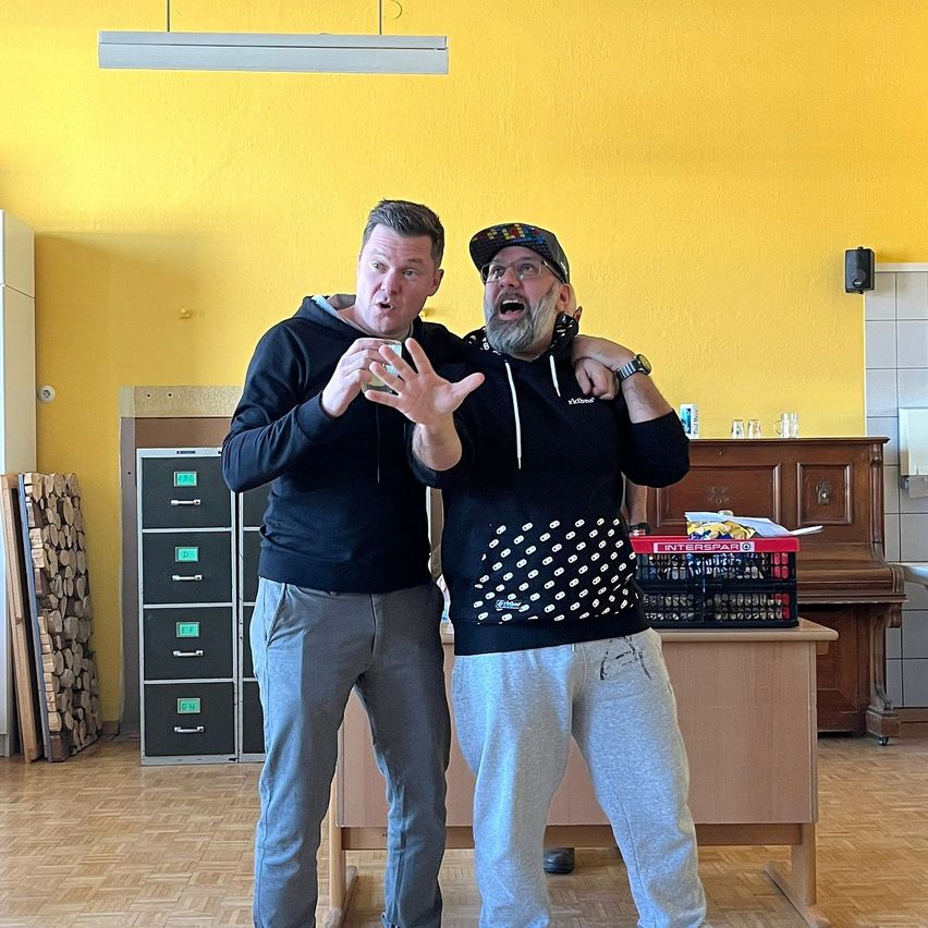 Two men stand in a room with yellow walls. One holds a phone and the other has his arm around the first man. Behind them is a wooden desk with a red crate and a piano.