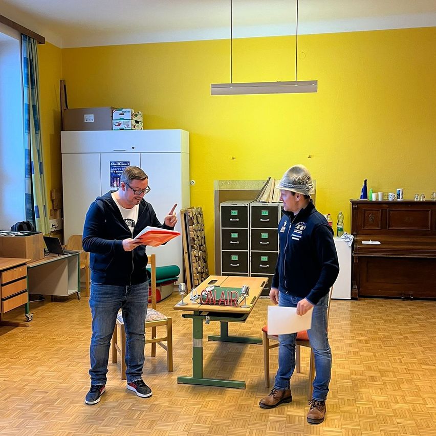 Two men stand in a room with yellow walls, holding papers. One man points at a book while the other holds a sheet of paper. Behind them, a table has a microphone and a sign reading 'ON AIR'. A cabinet and a piano are in the background.