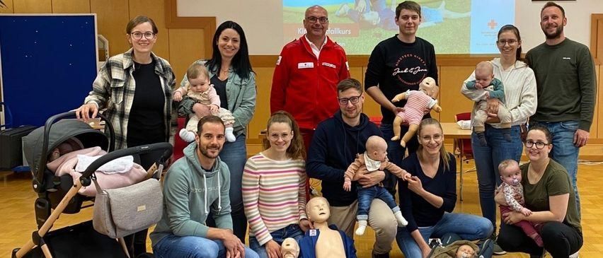 Eine Gruppe von Erwachsenen und Babys posiert für ein Foto in einem Raum. Hinter ihnen ist eine Projektionsleinwand und einige Personen tragen Brillen.