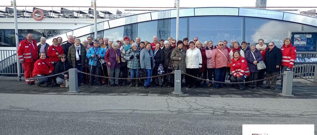 A group of people dressed in winter clothing pose for a photo in front of a building with banners. The photo has a watermark with text.